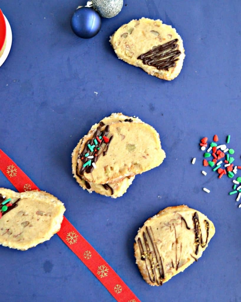 A blue background scattered with shortbread cookies that have been drizzled with chocolate and sprinkled with festive sprinkles. There's a red ribbon down the left side and sprinkles spilled on the right side. 