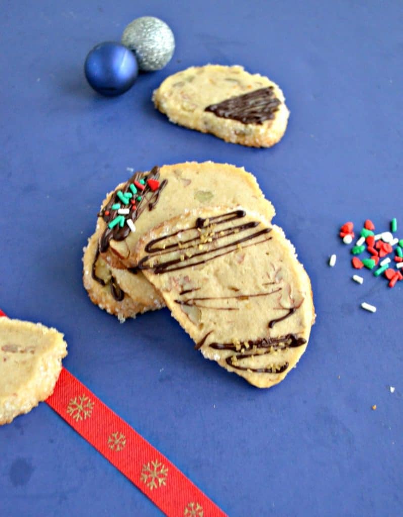 A close up of a shortbread cookie drizzled with chocolate leaning against a stack of cookie with another cookie in the background, spilled sprinkles to the right, and a red ribbon to the left. 