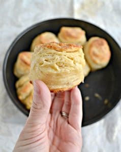 A close up view of a hand holding a fluffy, golden brown biscuit with a pan of biscuits in th background.