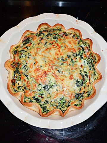 A pie pan with a Spinach Quiche that has a golden brown crust, the cheese on top has browned, and it's studded with spinach.