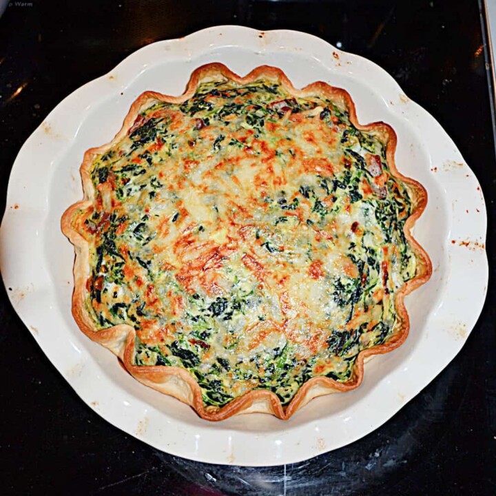A pie pan with a Spinach Quiche that has a golden brown crust, the cheese on top has browned, and it's studded with spinach.