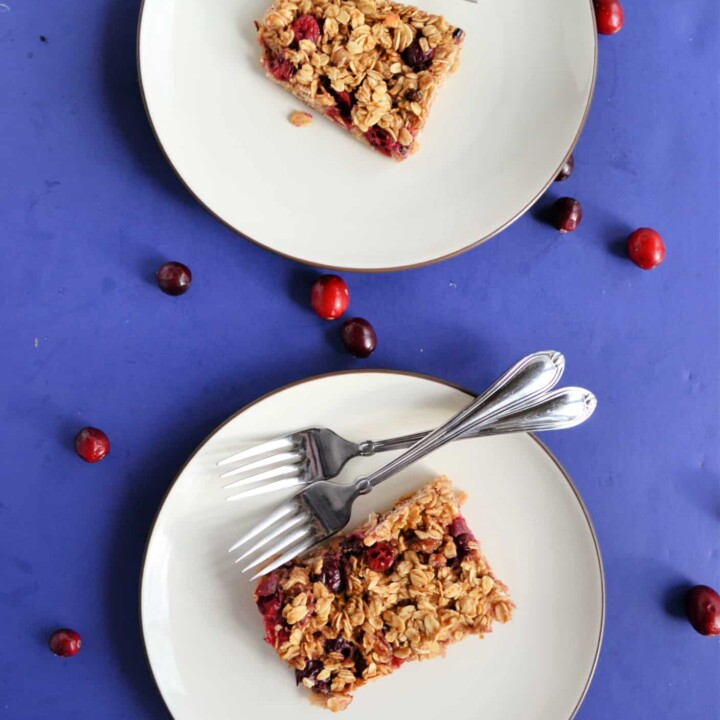 Two white plates each topped with a baked oatmeal square studded with red cranberries. The top plate has one fork on it and the bottom has two forks. There is a blue background with cranberries sprinkled on it.