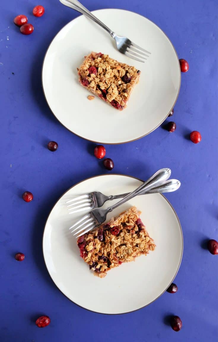 Two white plates each topped with a baked oatmeal square studded with red cranberries. The top plate has one fork on it and the bottom has two forks. There is a blue background with cranberries sprinkled on it.