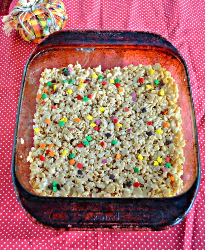 An overhead view of glass dish with Rice Krispies Treats sprinkles with colored leaf sprinkles on a red background and a scarecrow to the upper left. 
