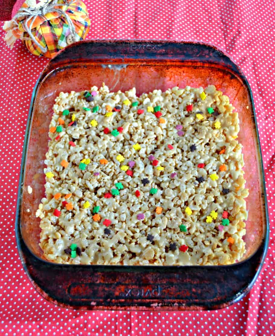 An overhead view of glass dish with Rice Krispies Treats sprinkles with colored leaf sprinkles on a red background and a scarecrow to the upper left.