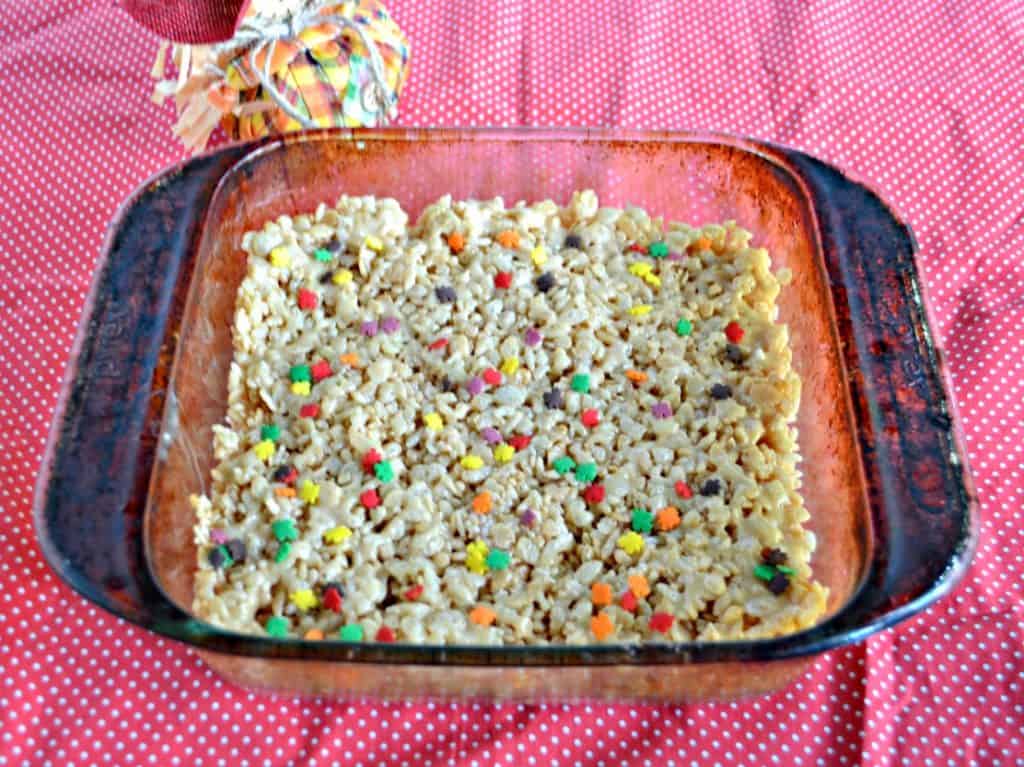 A glass dish with Rice Krispies Treats sprinkles with colored leaf sprinkles on a red background and a scarecrow to the upper left. 