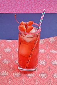 Cupid's Cocktail is a bright red cocktail in a tall glass rimmed with red sugar, two heart shaped strawberry garnishes, and a fun paper straw on a pink and blue background.