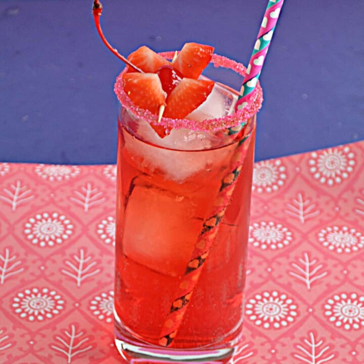 Cupid's Cocktail is a bright red cocktail in a tall glass rimmed with red sugar, two heart shaped strawberry garnishes, and a fun paper straw on a pink and blue background.