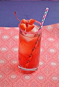 Cupid's Cocktail is a bright red cocktail in a tall glass over ice rimmed with red sugar, two heart shaped strawberry garnishes, and a fun paper straw on a pink and blue background.
