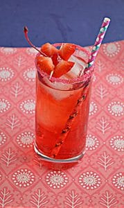 A tall glass filled with Cupid's Cocktail which is a bright red cocktail with a red sugared rim and two heart shaped strawberries and a paper straw on a pink background with a blue backdrop.