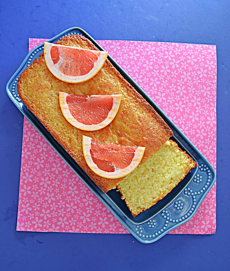 A loaf of grapefruit cake on a blue platter with three slices of grapefruit on top and a piece of the cake sliced off.