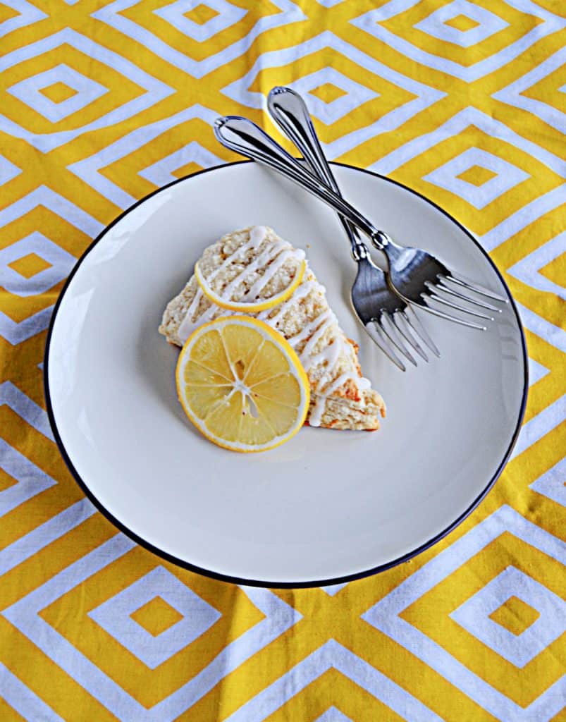 A large Meyer Lemon Scone on top of a white plate with a slice of lemon on top of it with two forks on the plate on a yellow and white background.