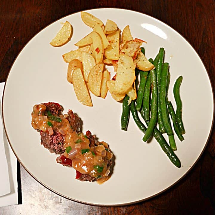 A white plate with a pile of steak fries, a pile of green beans, and a mini meatloaf sliced into four pieces with gravy on top.