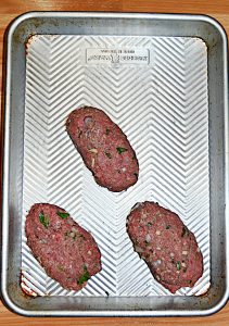 A silver sheet pan with three mini meatloaves shaped into logs on it.