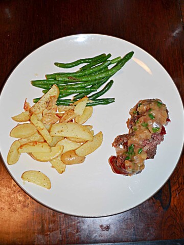 A white plate with a pile of steak fries, a pile of green beans, and a mini meatloaf sliced into four pieces with gravy on top.