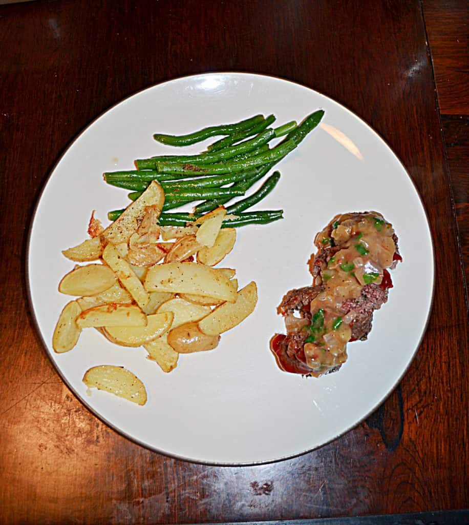A white plate with a pile of steak fries, a pile of green beans, and a mini meatloaf sliced into four pieces with gravy on top. 