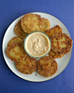 A white plate piled high with fried green tomatoes and a glass bowl of light orange remoulade sauce in the middle on a blue background.