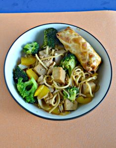 A large bowl filled with tofu, broccoli, and noodles with a golden brown eggroll in the upper right corner all on an orange background.