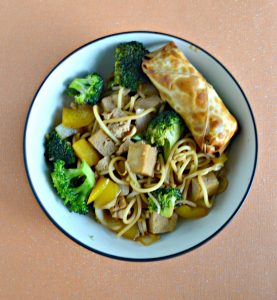 Overhead view of a large bowl filled with tofu, broccoli, and noodles with a golden brown eggroll in the upper right corner all on an orange background.