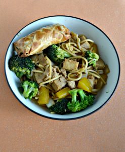 A large bowl filled with tofu, broccoli, and noodles with a golden brown eggroll in the upper left corner all on an orange background.