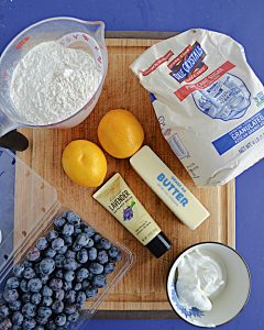 A cutting board topped with a cup of flour, a bag of sugar, 2 lemons, butter, lavender extract, a container of berries, and a bowl of sour cream.