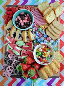 A large cutting board topped with colorful candies, cookies, quick breads, and nuts.