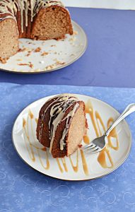 A plate with a slice of golden brown Bundt Cake topped with a glaze and the plate splatter with a caramel drizzle and the cut Bundt cake peeking in from the back left corner.