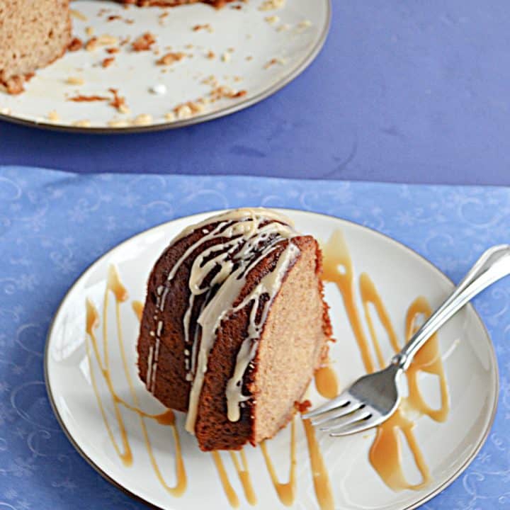 A plate with a slice of golden brown Bundt Cake topped with a glaze and the plate splatter with a caramel drizzle and the cut Bundt cake peeking in from the back left corner.