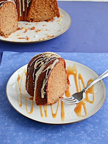 A close up of a plate with a slice of golden brown Bundt Cake topped with a glaze and the plate splatter with a caramel drizzle and the cut Bundt cake peeking in from the back left corner.