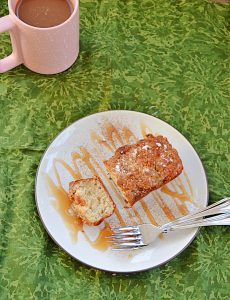 A top view of a plate drizzled with caramel sauce with a mini pear ginger cake on it and two forks on the plate with a cup of coffee in the background.