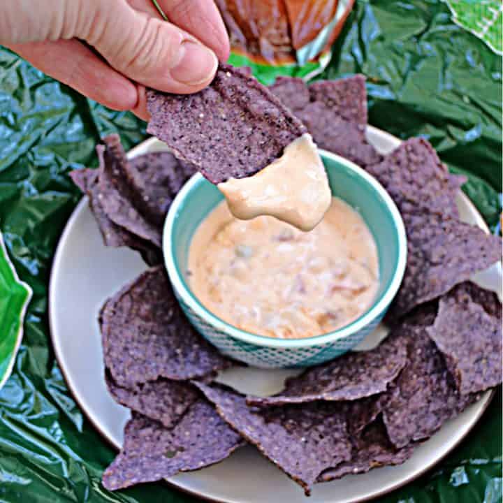 A plate of blue tortilla chips with a bowl of creamy queso in the middle and a hand dipping a chip into the queso.