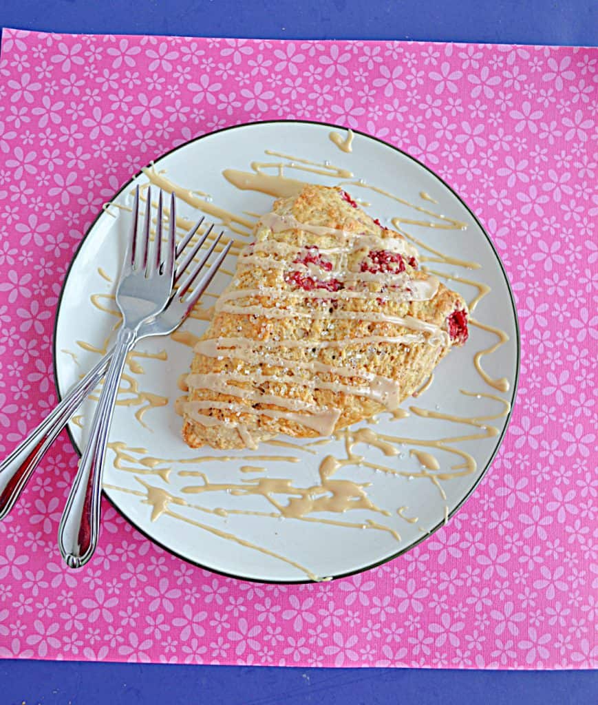 A plate with a raspberry almond scone drizzled with coffee glaze and two forks on the plate with a pink background.