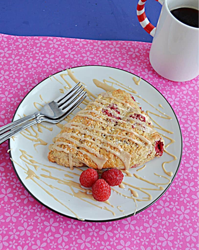 A plate with a raspberry almond scone drizzled with coffee glaze and two forks on the plate along with 3 raspberries. There's a cup of coffee behind the scone.