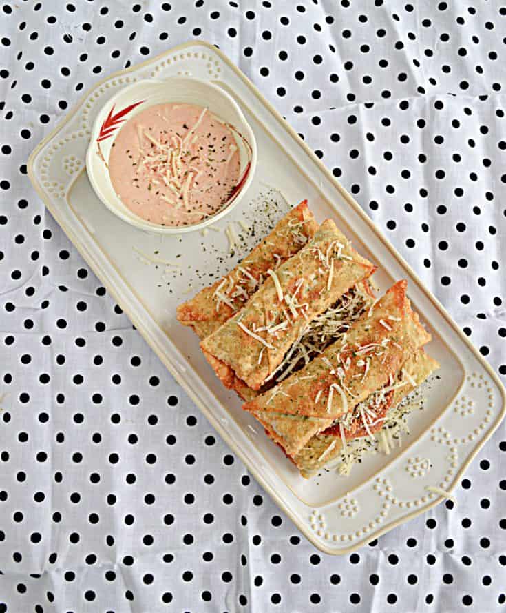 A white platter piled high with crispy egg rolls and served with a small white bowl of spicy dipping sauce on a black and white polka dot background.