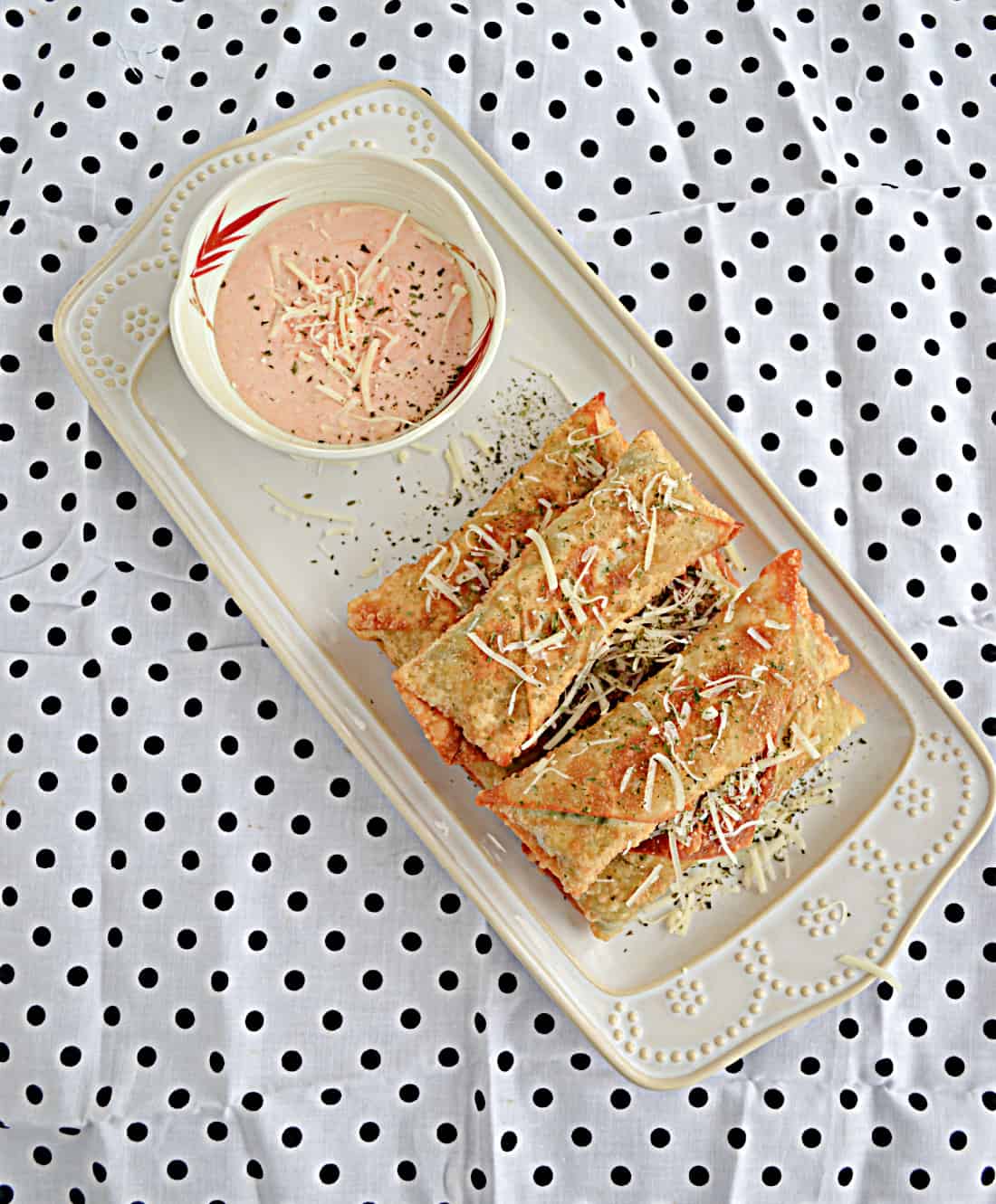 A white platter piled high with crispy egg rolls and served with a small white bowl of spicy dipping sauce on a black and white polka dot background.