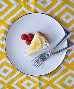 A plate topped with a sliced of lemon lavender cheesecake with a lemon slice on top, three raspberries on the side, and two forks on the plate.