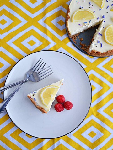 A top view of A plate topped with a sliced of lemon lavender cheesecake with a lemon slice on top, three raspberries on the side, and two forks on the plate with the cut cheesecake in the background.