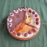 A cheesecake divided into 3 sections: One is orange cranberry with whole cranberries on top, one third is caramel pecan cheesecake topped with caramel and pecan halves, and one third is chocolate espresso topped with espresso beans.