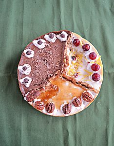 A top view of a cheesecake divided into 3 sections: One is orange cranberry with whole cranberries on top, one third is caramel pecan cheesecake topped with caramel and pecan halves, and one third is chocolate espresso topped with espresso beans.