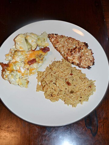 A top view of a plate topped with a crispy almond coated chicken breast, a pile of rice, and loaded cauliflower