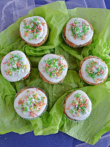 Top view of seven carrot cake cupcakes topped with cream cheese frosting and sprinkles on a green background.