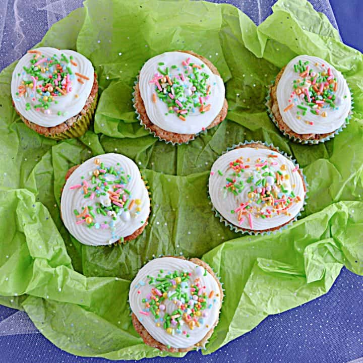 A pile of carrot cake cupcakes topped with cream cheese frosting and sprinkles on a green background.