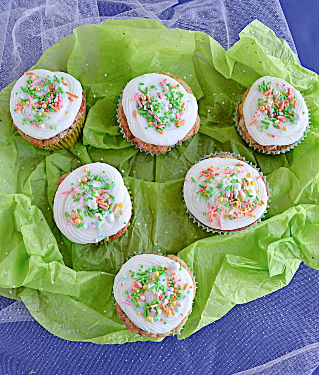 A pile of carrot cake cupcakes topped with cream cheese frosting and sprinkles on a green background.