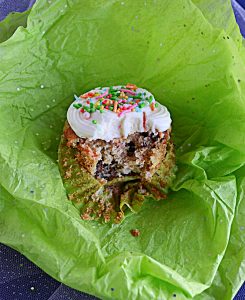 A carrot cake cupcake topped with cream cheese frosting and sprinkles with a big bite taken out of it sittin gon green tissue paper.