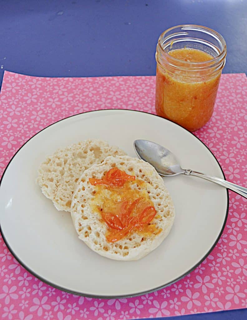 A plate with an English Muffin topped with Kumquat jam with a jar of orange Kumquat Jam behind the plate.
