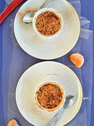 A top view of two plates with two ramekins on them with golden brown crumble on top each with two spoons on the plate with a stalk of rhubarb at the top and half an orange beside the plates.