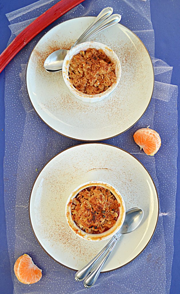 A top view of two plates with two ramekins on them with golden brown crumble on top each with two spoons on the plate with a stalk of rhubarb at the top and half an orange beside the plates. 