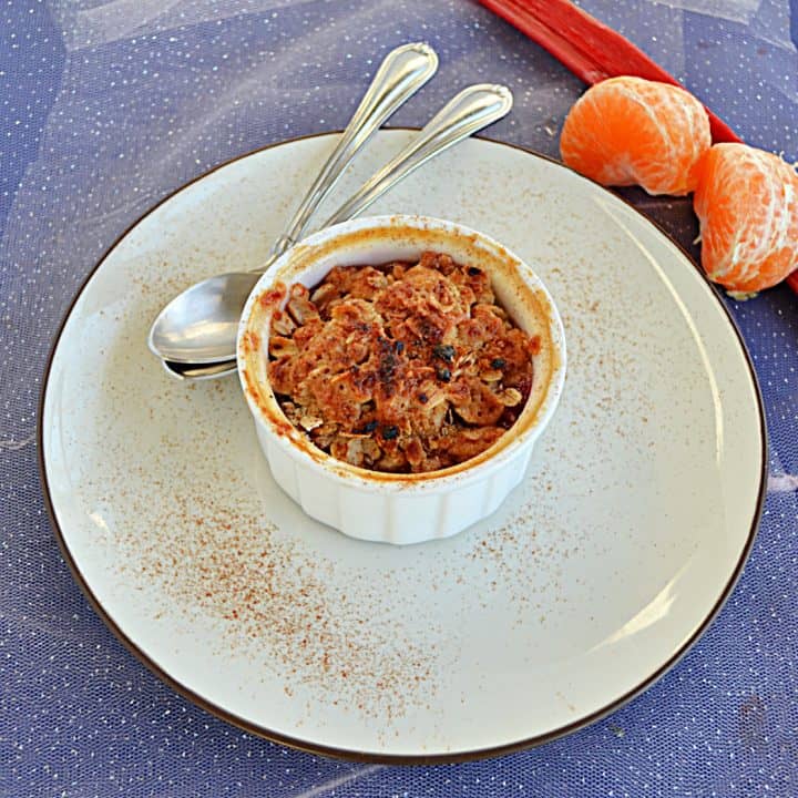 A ramekin of Orange Rhubarb Crisp topped with golden brown crumble on a plate with two spoons and a stalk of rhubarb with two oranges behind the plate.