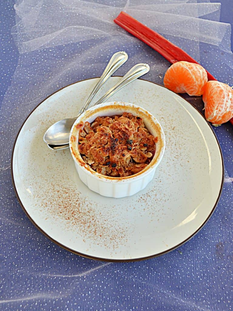 A ramekin of Orange Rhubarb Crisp topped with golden brown crumble on a plate with two spoons and a stalk of rhubarb with two oranges behind the plate. 