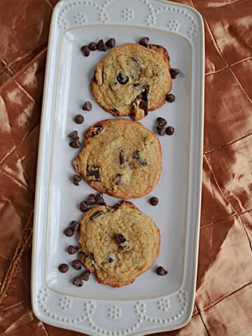 A platter with three chocolate chip cookies on it and chocolate chips sprinkled over top.